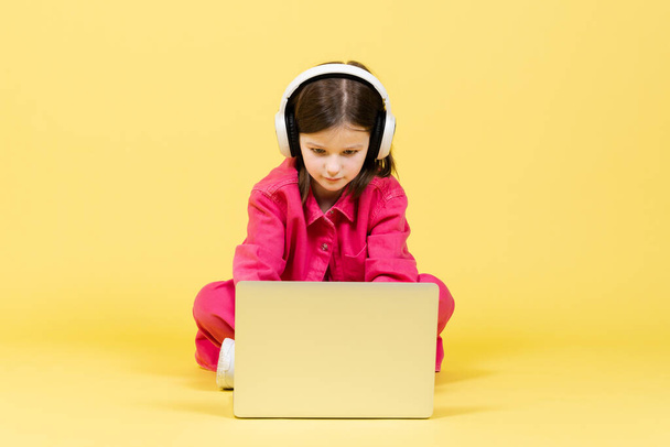かわいい女の子が床に座ってラップトップを使用しています。コンピュータに集中してヘッドフォンを身に着けている人形. - 写真・画像