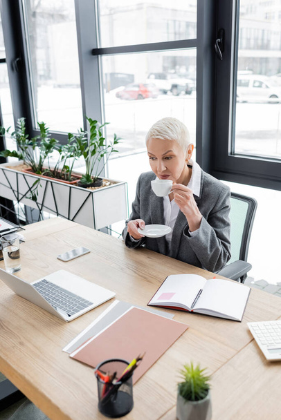 senior economist drinking coffee at workplace near laptop and blank notebook - Zdjęcie, obraz