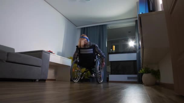Młody człowiek na wózku inwalidzkim słucha muzyki w słuchawkach i kręci się po pokoju - Materiał filmowy, wideo
