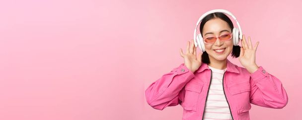 Танцююча стильна азіатська дівчина слухає музику в навушниках, позує на рожевому фоні
 - Фото, зображення
