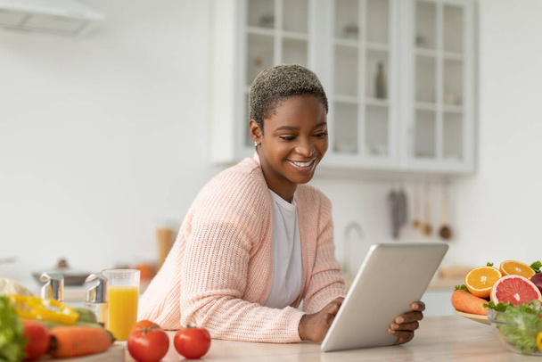 Усміхаючись тисячоліття афроамериканська леді блогер дивиться на планшет за столом з органічними фруктами
 - Фото, зображення
