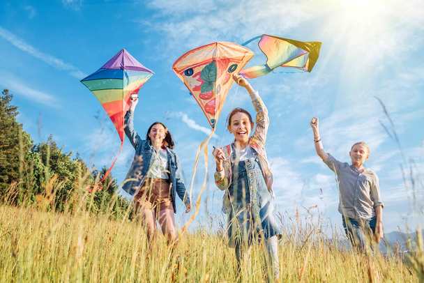 Smiling gils and brother boy running with flying colorful kites on the high grass meadow in the mountain fields. Happy childhood moments or outdoor time spending concept image. - Foto, immagini