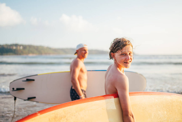 Smiling teen boy with surfboard go to the sea for surfing. He have a winter vacation and enjoying a beautiful sunset light with father on Sri Lanka island. Family active vacation concept.  - Foto, Imagem