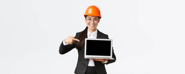 Gülümseyen Asyalı kadın baş mühendis şirket karıyla grafiği gösteriyor, yatırımcılar toplantısında sunum yapıyor, güvenlik kaskı takıyor, dizüstü bilgisayara işaret ediyor, beyaz arka plan - Fotoğraf, Görsel