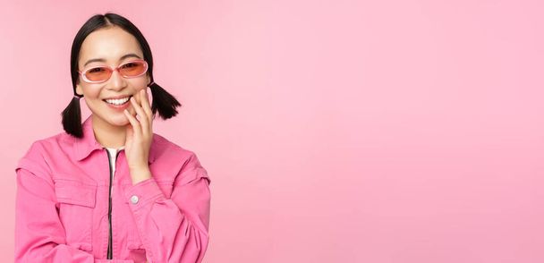 Close up ritratto di elegante donna asiatica in occhiali da sole, sorridente, cercando lusingato, ridendo civettuola, in piedi su sfondo rosa - Foto, immagini