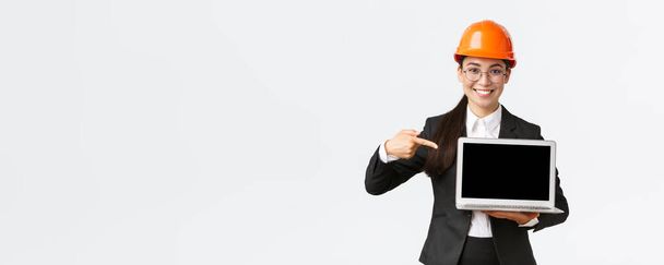 Smiling pleased asian female chief engineer showing chart with enterprise profit, making presentation during investors meeting, wearing safety helmet, pointing at laptop screen, white background - Foto, imagen