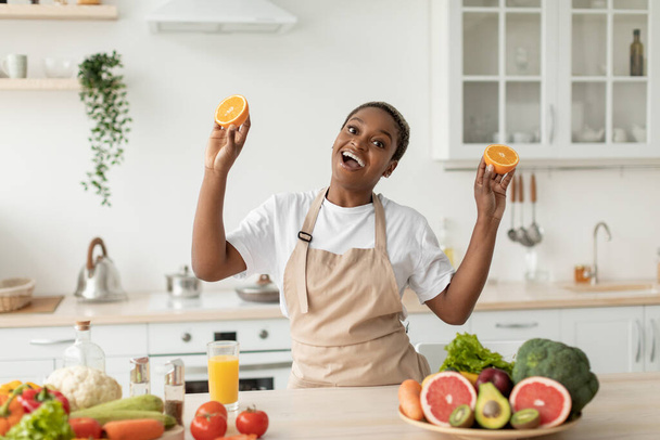 Feliz emocional jovem mulher negra no avental se divertir no interior da cozinha minimalista, mantém laranjas - Foto, Imagem