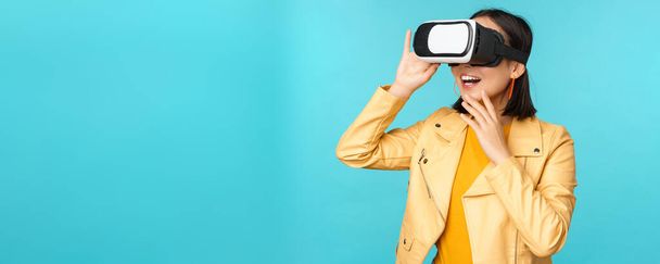Portrait of happy asian female model using VR headset, smiling and laughing in virtual reality glasses, standing over blue background - Photo, Image