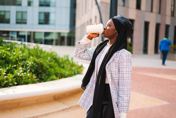 Black woman in Islamic scarf drinking coffee in city park - Photo, Image