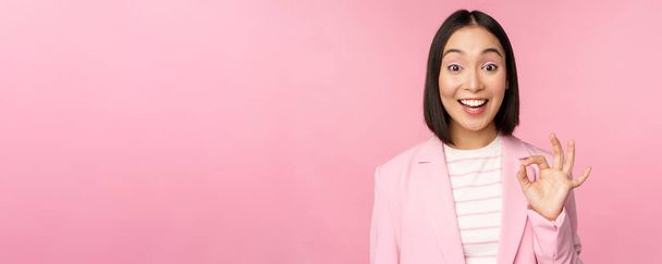 Very good. Happy korean businesswoman smiling pleased, showing okay, ok sign, well done, excellent choice gesture, praise smth, standing over pink background - Photo, image