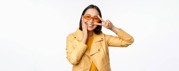 Portret stylowej azjatyckiej nowoczesnej dziewczyny, ubranej w okulary przeciwsłoneczne i żółtą kurtkę, pokazujący spokój, gest migowy, stojący nad białym tłem, szczęśliwa uśmiechnięta twarz - Zdjęcie, obraz