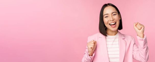 Захоплена продавчиня, азіатська корпоративна жінка каже "так", досягає мети і святкує, тріумфує, дивлячись з радістю і посмішкою, стоїть над рожевим тлом
 - Фото, зображення