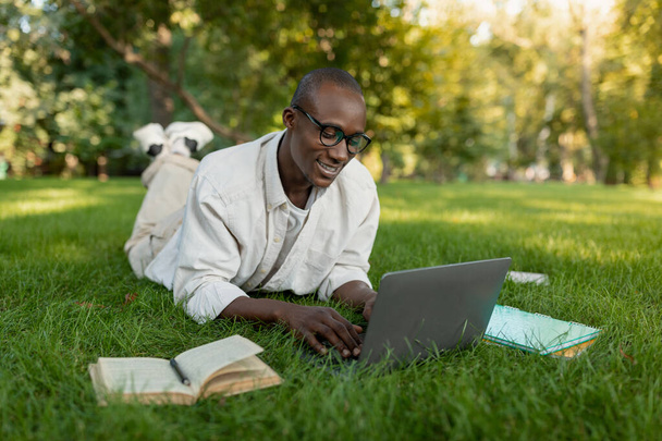 Cheerful black student guy using laptop while learning online outdoors, lying on grass in college campus - Valokuva, kuva