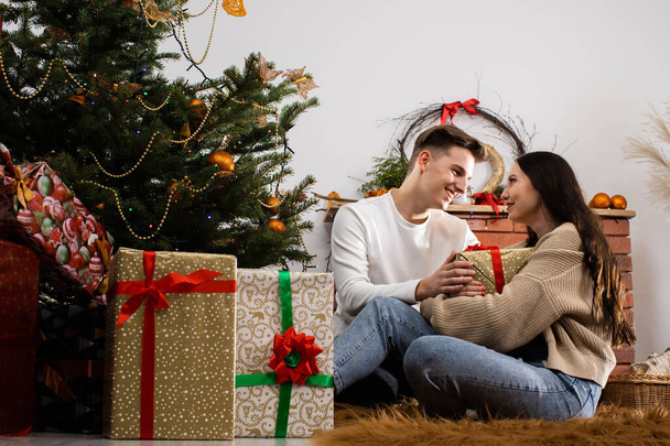Viewing gift packages. Couple in love with a gift sitting on the floor by the Christmas tree on Christmas. A unique Christmas atmosphere. - Фото, зображення