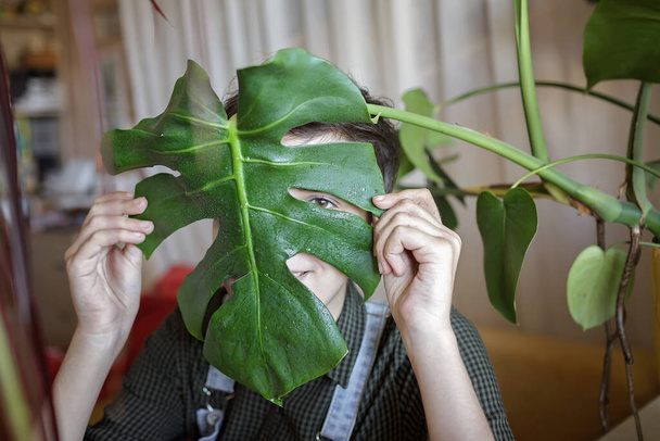 若い女の子は楽しさとリラックスのために緑の葉で彼女の顔をカバーします,バルコニーの自宅の植物, gen Z - 写真・画像