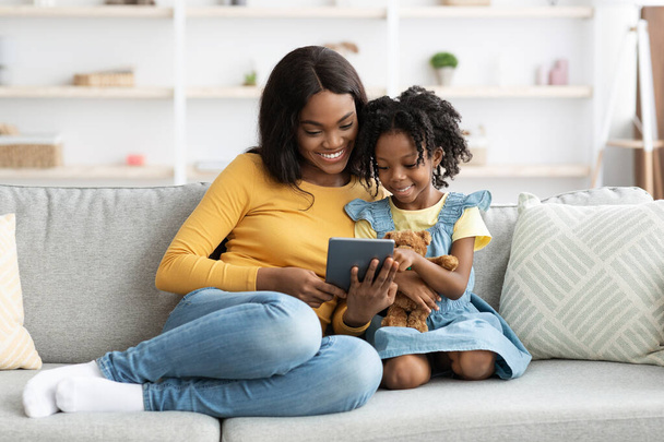 陽気な若いアフリカ系アメリカ人のお母さんと小さな娘は、デジタルタブレットでリラックス - 写真・画像
