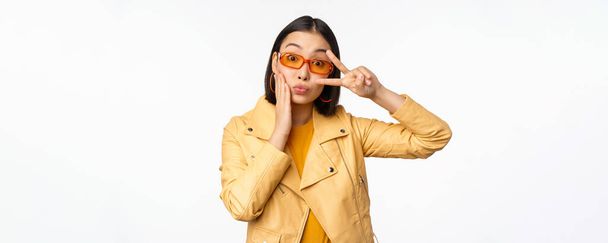 Retrato de menina moderna asiática elegante, vestindo óculos de sol e casaco amarelo, mostrando paz, gesto v-sign, de pé sobre fundo branco, rosto sorridente feliz - Foto, Imagem