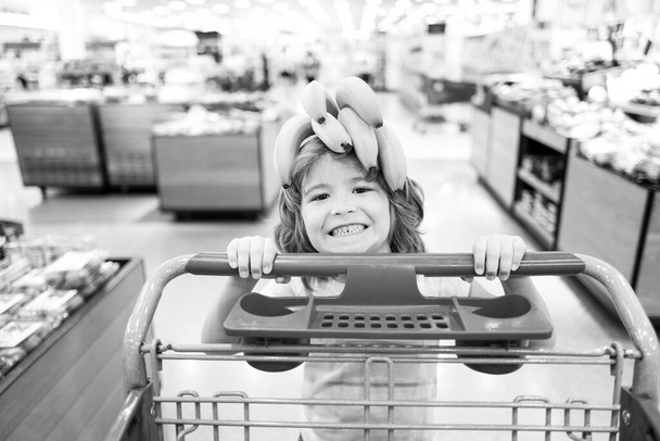Child with shopping cart at grocery store or supermarket. Kid buying banana fruit in grocery market. - Фото, изображение