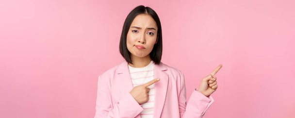 Скептически настроенная азиатская бизнесвумен, продавщица дуется и смотрит с неодобрением, указывая пальцем направо, показывая плохую информацию, расстраивая новости, стоя на розовом фоне - Фото, изображение