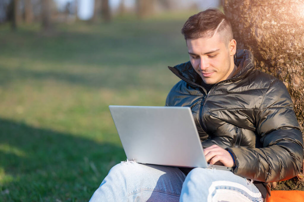 Összpontosított jóképű férfi alkalmi viseletben, laptoppal dolgozik a szabadban. Egy fa lábánál ülve élvezi a természetet egy napos tavaszi napon. Távmunka koncepció. Fénymásolási hely. - Fotó, kép