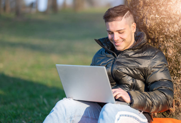 Homem bonito focado no desgaste casual que trabalha com laptop ao ar livre. Sentado ao pé de uma árvore, ele gosta da natureza em um dia ensolarado de primavera. Conceito de trabalho remoto. Espaço de cópia. - Foto, Imagem