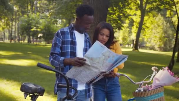 Jong Afrikaans-Amerikaans koppel bestudeert kaart, plant route door park, geniet van fietstocht, tracking shot - Video