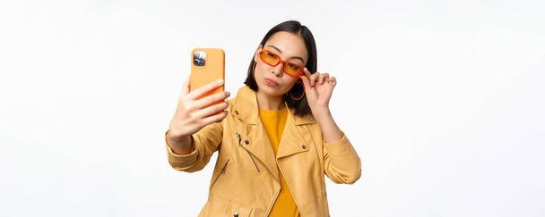 Stylowa, piękna azjatycka dziewczyna w okularach przeciwsłonecznych, robienie selfie na smartfonie, pozowanie do zdjęcia, trzymanie telefonu komórkowego, białe tło studio - Zdjęcie, obraz