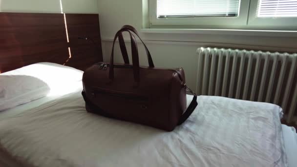 Bolsa de cuero marrón se sienta en la cama individual del hotel en la luz de la mañana desde la ventana - Metraje, vídeo