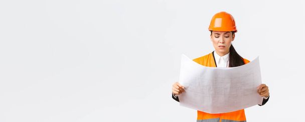 Confused and doubtful asian female chief engineer, lead architect at construction area looking displeased and hesitant at strange blueprints, reading confusing building project, white background - Photo, Image