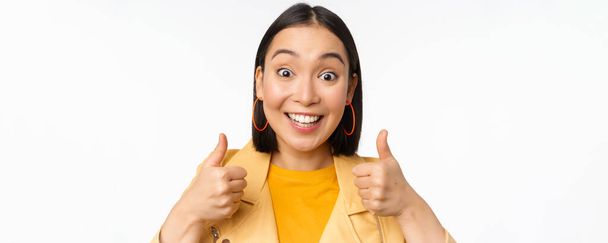 Sonriendo chica asiática mostrando los pulgares hacia arriba, mirando complacido, aprobar smth, de pie sobre fondo blanco - Foto, imagen
