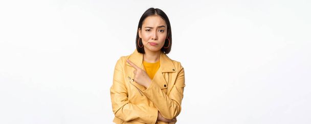 Portrait of stylish asian girl looking with doubt, skeptical face, pointing finger left at logo or advertisement, standing against white background - Photo, image