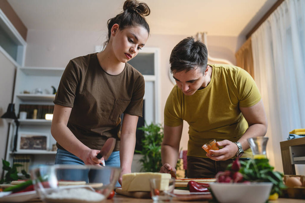 Two people young adult couple man and woman husband and wife or boyfriend and girlfriend preparing pizza food at home helping each other cutting and add ingredients and spices real people copy space - Photo, Image