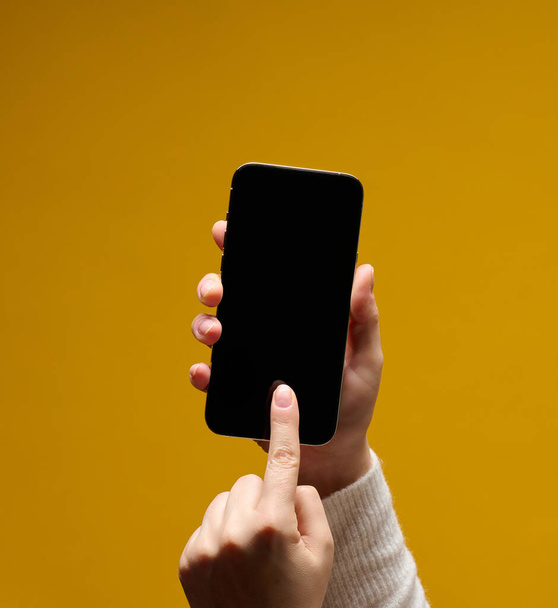 女性の手は黄色の背景に黒い画面でスマートフォンを保持 - 写真・画像