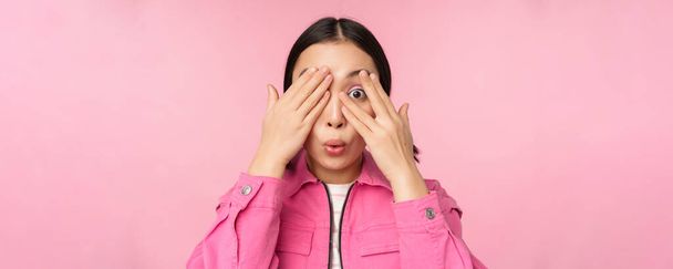 Close up portrait of young asian girl looking surprised, express amazement and wonder, peeking through fingers, standing over pink background - Photo, Image