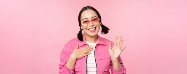 Πορτρέτο της φιλικής Ασιάτισσας έφηβης με γυαλιά ηλίου που λέει γεια, κουνάει το χέρι της και χαμογελάει, σε χαιρετά, γεια χειρονομία, στέκεται πάνω από ροζ φόντο - Φωτογραφία, εικόνα