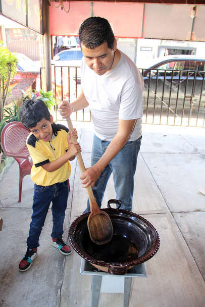 Latin dad and son cook together the traditional Mexican dish mole rojo in a clay pot with a wooden spoon as a family activity of pride, culture and tradition - Photo, Image