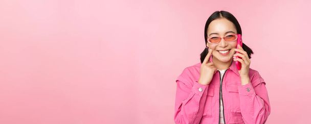 Красивая корейская модель в солнцезащитных очках, разговаривающая по мобильному телефону со счастливым лицом, использующая сотовую связь, чтобы позвонить другу по смартфону, стоя на розовом фоне - Фото, изображение