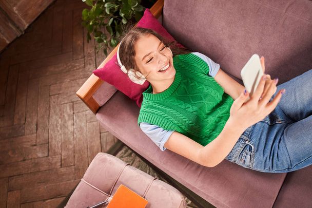 Cheerful teenager girl wearing headphones laying at the sofa and listening music - Photo, image