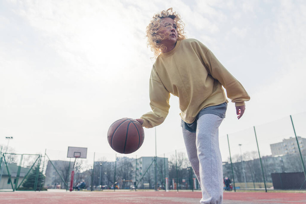 Attractive blond woman plays basketball in baggy clothes, looking up copy space medium full shot  - Фото, изображение