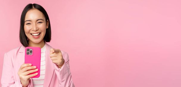 Feliz asiática mujer de negocios riendo, señalando con el dedo en usted y la grabación de vídeo, tomando fotos en el teléfono inteligente, utilizando el teléfono móvil, fondo rosa - Foto, imagen