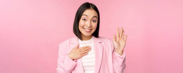 Retrato de linda mujer de negocios asiática levantando la mano, presentarse en la oficina, sonriente coqueta, de pie sobre fondo rosa en traje - Foto, imagen