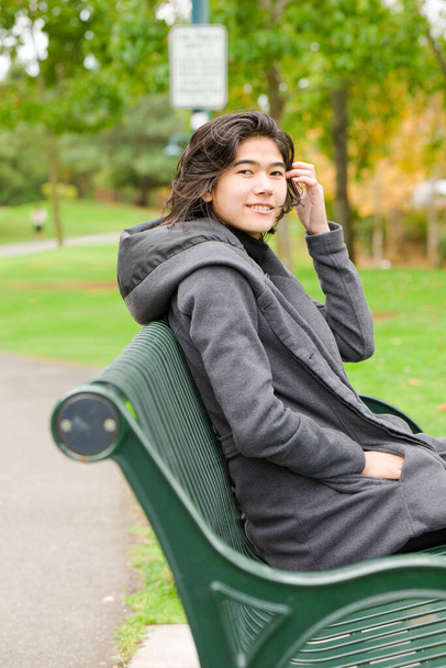Smiling biracial teen girl or young adult female in gray jacket sitting on green park bench by lake on overcast autumn or spring day - Foto, immagini
