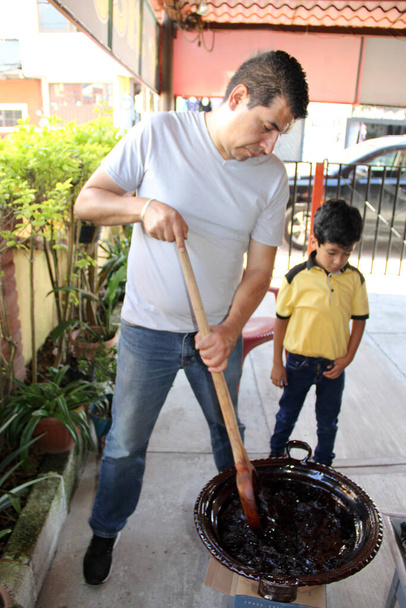 Pai e filho latino cozinham juntos o tradicional prato mexicano mole rojo em uma panela de barro com uma colher de madeira como uma atividade familiar de orgulho, cultura e tradição. - Foto, Imagem