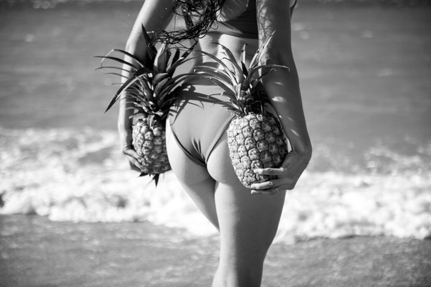 Female closeup buttocks in thongs bikini, sexy ass. Young woman holding a pineapple on sea sand beach background. - Foto, Imagen