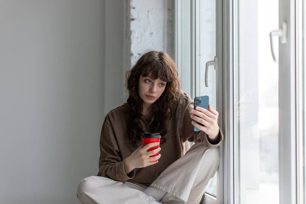Young attractive woman with a cup of coffee in her hands makes a selfie sitting on the windowsill. - Φωτογραφία, εικόνα