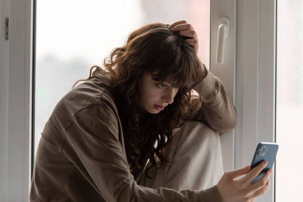 Young attractive woman uses smartphone while sitting on the windowsill by the window. - Фото, зображення