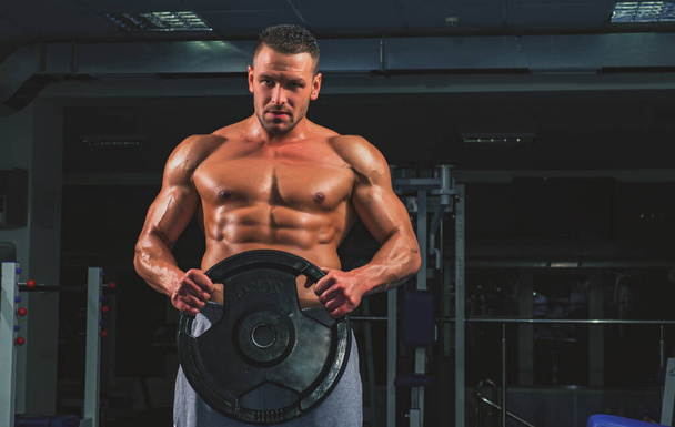 Sport Gym. Sportsman with weight disk in gym. Bodybuilder with heavy disk trains biceps. - Zdjęcie, obraz