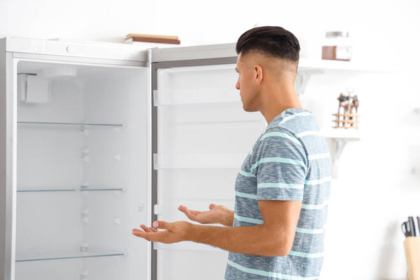 Запутавшийся человек рядом с пустым холодильником на кухне - Фото, изображение