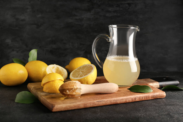 Wooden board with ripe lemons, juicer and jug of juice on dark background - Fotó, kép