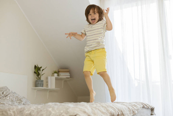 A little boy joyfully jumps on the bed in the bedroom. Fun games at home. Childrens activity and games with parents - Фото, изображение
